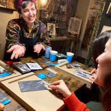 Load image into Gallery viewer, A linoprint workshop in a cosy, warm shop. The teacher is smiling at the student - explaining how to carve or mark make