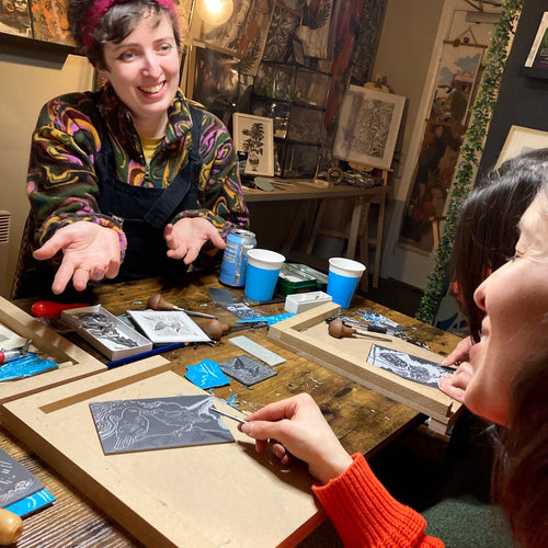 A linoprint workshop in a cosy, warm shop. The teacher is smiling at the student - explaining how to carve or mark make