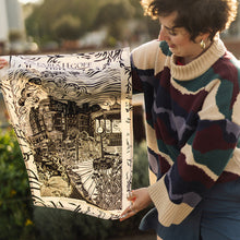 Load image into Gallery viewer, Person holding a linocut teatowel outdoors with greenery and a building in the background
