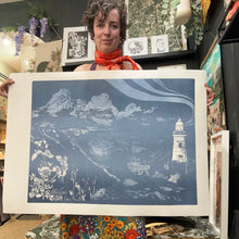 Load image into Gallery viewer, A person in a shop holds up a large square art print featuring a blue and white illustration of a coastal landscape with clouds, a lighthouse, and waves. The print has a wide white border, and the background shows framed artwork and shelves in a gallery or studio setting.

